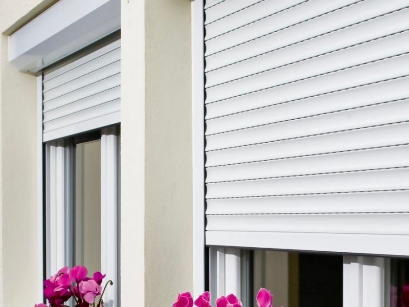 Exterior view of windows with closed white blinds and a pot of pink flowers on the windowsill.